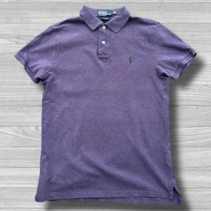 Ralph Lauren piké - Säljer en stilren lila pikétröja från Ralph Lauren. Tröjan har en klassisk krage med två knappar och det ikoniska gröna broderade logotypen på bröstet. Perfekt för en avslappnad men ändå elegant look. Passar bra till både jeans och chinos.