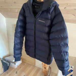 Jacka  - Säljer en snygg mörkblå dunjacka från Peak Performance. Jackan har en dragkedja framtill och en praktisk huva. Perfekt för kyliga dagar med sin pufferdesign som håller dig varm och bekväm.