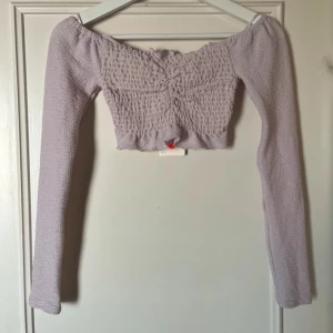 Ljuslila offshoulder topp från BikBok - Säljer en söt ljuslila offshoulder topp från BikBok. Toppen är croppad med långa ärmar och har en smockad design som ger en fin passform. Perfekt för att lägga till en romantisk touch till din outfit! 