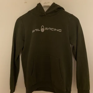 Grön hoodie från Sail Racing - Säljer en snygg grön hoodie från Sail Racing med tryck på bröstet. Tröjan har en klassisk känguruficka och långa ärmar. Perfekt för en avslappnad stil.