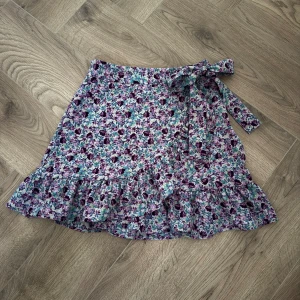 Blommig omlottkjol med volang - Säljer en söt blommig omlottkjol med volangdetaljer och knytband i midjan. Kjolen har en härlig mix av lila, blå och gröna nyanser. Perfekt för en somrig look!