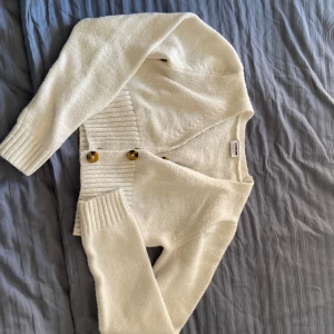 Vit kofta från Noisy May - Säljer en stilren vit kofta från Noisy May. Den har långa ärmar och 3 stora knappar framtill. Perfekt för att hålla sig varm och snygg under kyligare dagar. Koftan har en ribbad kant vid ärmslut och nederkant.