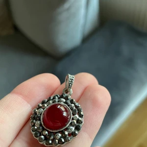 Hänge med röd sten - Elegant hänge med en stor röd sten i mitten, omgiven av en dekorativ silverfärgad ram. Perfekt som ett statement-smycke för att lyfta vilken outfit som helst.
