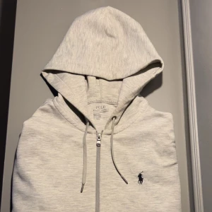 Grå hoodie från Polo Ralph Lauren - Säljer en stilren grå hoodie från Polo Ralph Lauren med dragkedja och justerbar huva. STORLEK M
