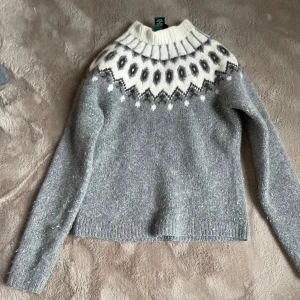 Grå stickad tröja från Ralph Lauren - Säljer en exklusiv handstickad grå tröja från Ralph Lauren med ett vackert mönster i vitt och svart runt halsen. Tröjan har långa ärmar och en ribbad krage. Perfekt för kyliga dagar!