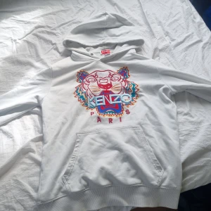 Vit hoodie från Kenzo - Säljer en vit hoodie från Kenzo med ett färgglatt tigerbroderi på framsidan. Hoodien har en klassisk passform med en stor ficka framtill och en bekväm huva. Perfekt för en stilren och trendig look.