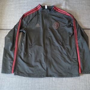 Svart träningsjacka från Adidas - Snygg svart träningsjacka från Adidas med röda ränder längs ärmarna och dragkedja framtill. Jackan har två fickor med dragkedjor och ett emblem på bröstet. Finns även två fickor på insidan men har hål i båda 