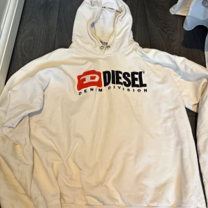 Vit hoodie från Diesel - Säljer en vit hoodie från Diesel med stor logga på framsidan i svart och rött. Hoodien har en klassisk design med huva och ribbade muddar. Perfekt för en avslappnad stil.