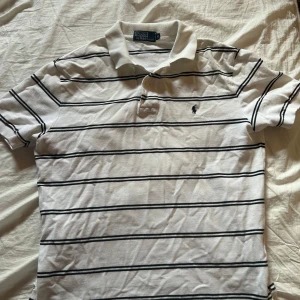 Vit och svart randig pikétröja från Ralph Lauren - Säljer en klassisk vit pikétröja från Ralph Lauren med svarta ränder. Tröjan har korta ärmar och en broderad logga på bröstet. Perfekt för en stilren look!