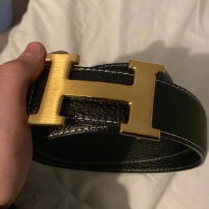 Svart skinnbälte med guldspänne - Snyggt svart skinnbälte med ett stort, iögonfallande guldspänne format som ett 'H'. Perfekt för att ge en stilren touch till din outfit. Passar både till jeans och kostymbyxor. Ni får box, kort och en påse till det 