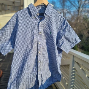 Blå och vit rutig skjorta från Ralph Lauren - Säljer en klassisk blå och vit rutig skjorta från Ralph Lauren. Skjortan har korta ärmar och knappar framtill. Den är broderad med den ikoniska loggan i grönt på bröstet. Perfekt för en stilren look!