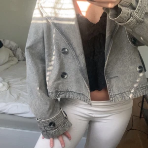 Grå croppad jeansjacka - Säljer en snygg grå croppad jeansjacka med stora knappar. Jackan har en cool, avslappnad stil och är perfekt för en trendig look. Den har långa ärmar och en klassisk krage. Från Gina tricort 