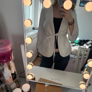 Vit kavaj med knytband - Snygg vit kavaj med knytband i midjan för en elegant look. Den har långa ärmar och en stilren design utan krage. Perfekt för att lyfta vilken outfit som helst.
