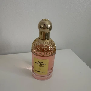 Aqua Allegoria Rosa Rossa från Guerlain - Elegant parfymflaska med rosa vätska och ett gyllene bikakemönster på korken. Aqua Allegoria Rosa Rossa är en delikat doft från Guerlain, perfekt för den som älskar blommiga och fruktiga toner. Flaskan har en lyxig design med en rundad gyllene kork.
