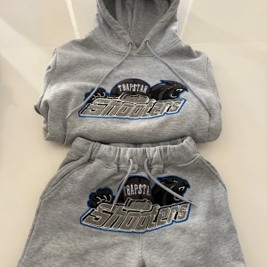 Grå set från Trapstar - Säljer en grå set från Trapstar med ett coolt 'Shooters' tryck i svart och blått på framsidan. Settet har en klassisk passform med dragsko i huvan och ribbade muddar vid ärmslut och nederkant. Perfekt för en avslappnad stil.