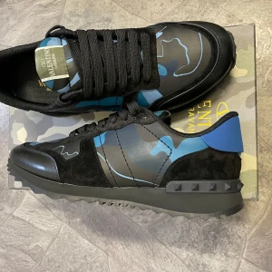 Valentino Rockrunner sneakers i blått och svart - Snygga Valentino Rockrunner sneakers med ett coolt blått och svart camouflagemönster. Skorna har svarta snören och en robust sula med nitar på hälen. Perfekta för att ge din outfit en edgy touch. Den passar folk med storlek mella 44-42