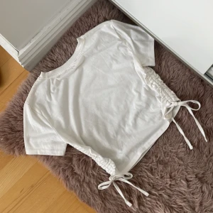 Vit croppad t-shirt med snörning i sidorna - Säljer en jätte fin vit croppad t-shirt med korta ärmar och snörning i båda sidorna. Toppen har en enkel och stilren design med rund halsringning och justerbara knytband i midjan. Står inte märke eller storlek men skulle säga xs/s. Hör av dig om du har grå eller funderingar 💕