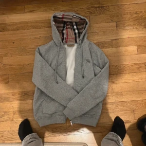 Grå hoodie från Burberry med klassiskt rutigt foder - Säljer en grå hoodie från Burberry med dragkedja och ikoniskt rutigt foder i huvan. Tröjan har långa ärmar, ribbade muddar och är tillverkad i mjuk bomull. Perfekt för en stilren och bekväm look.