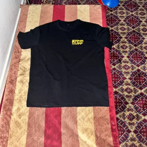Svart t-shirt med färgglatt tryck från Sana Duri - Svart t-shirt med coolt och färgglatt tryck på ryggen med svamp, öga och texten Sana Duri. Liten gul logga på bröstet. Perfekt för dig som gillar unika och kreativa plagg.