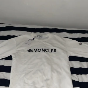 Vit sweatshirt från Moncler - Säljer en vit sweatshirt från Moncler med svart logotyptryck på bröstet och klassisk Moncler-patch på ärmen. Tröjan har rund halsringning och långa ärmar.