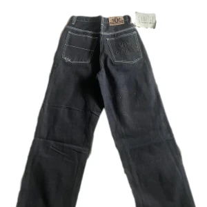 Svarta jeansbyxor från Outkast med broderad ficka - Säljer ett par svarta jeansbyxor från Outkast med vit kontrastsöm och broderad logga på bakfickan. Klassisk femficksmodell med raka ben och normal passform. Snygga detaljer och läderpatch i midjan.