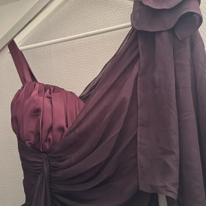 Vinröd långklänning med drapering och rosett - Säljer en vinröd långklänning med ett axelband och vacker drapering över hela klänningen. Klänningen har en stor rosett på axeln och är tillverkad i ett glansigt, tunt material som ger ett elegant intryck.