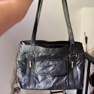 Svart axelväska i skinnimitation - Snygg svart axelväska med flera fack och dragkedjor framtill och baktill. Väskan har långa handtag och rymligt huvudfack med innerficka. Perfekt för dig som vill ha plats för allt du behöver i vardagen.