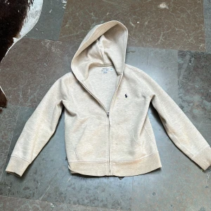 Beige hoodie med dragkedja från Polo Ralph Lauren - Säljer en beige hoodie från Polo Ralph Lauren i storlek M (10-12). Tröjan har dragkedja framtill, huva och den klassiska broderade loggan på bröstet. Perfekt för en avslappnad och stilren look.Köpt i USA på Ralph Lauren butik.