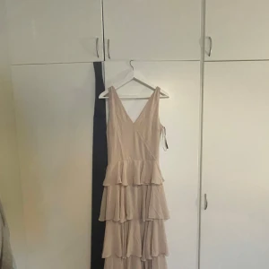 klänning med volanger - Säljer en elegant gräddvit långklänning med flera lager volanger och v-ringning från ASOS. Klänningen är ärmlös och har en feminin siluett som passar perfekt till speciella tillfällen. Den har aldrig blivit använd och den är sydd vid bröstet då det var för öppet.