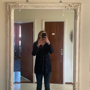 Mörkblå parkajacka med huva - Säljer en mörkblå parkajacka med huva och dragsko i midjan. Jackan har dragkedja framtill, stora fickor med lock och långa ärmar. Perfekt för blåsiga dagar och enkel att matcha med olika outfits.