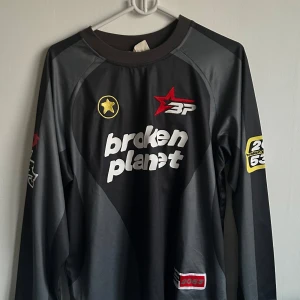 Svart långärmad tröja från Broken Planet - Cool svart långärmad tröja från Broken Planet med tryck på både fram- och baksida. Tröjan har sportiga detaljer, vita och gula loggor samt nummer 12 på ryggen. Perfekt för dig som gillar streetwear och unika plagg.
