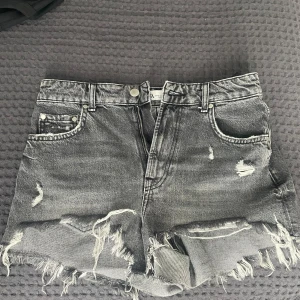 Grå jeansshorts från ZARA - Säljer ett par grå denimshorts från ZARA med slitna detaljer. Shortsen har hög midja och dragkedja med knapp. Shortsen är knappt använda och därför i nyskick. 
