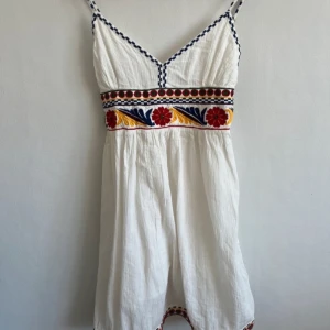 Vit klänning med broderade detaljer - Superfin vit klänning med smala axelband och färgglada broderier i rött, blått och gult runt midjan och längs kanterna. Klänningen har v-ringning och är perfekt för varma dagar.