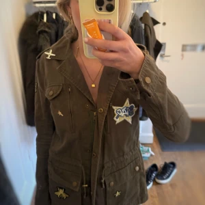 Olivgrön militärinspirerad jacka med patches - Säljer en olivgrön militärjacka från Blue Monkey  med flera coola patches, bland annat stjärna, bi och fjäder på ryggen. Jackan har flera fickor, knappar och dragkedja framtill samt dekorativa detaljer på axlar och bröst. Knappt använd.