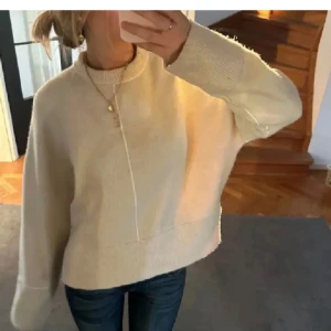 Beige stickad tröja från Zara - Säljer en beige stickad tröja med hög hals och långa ärmar. Tröjan har en avslappnad passform och synlig söm framtill som detalj. Perfekt för kyligare dagar och enkel att matcha med jeans eller kjol.