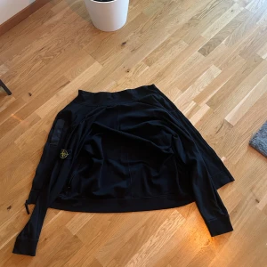 Svart zip up från Stone Island - Säljer en svart jacka från Stone Island med klassisk patch på ärmen. Jackan har lång ärm och en stilren design som passar till många tillfällen. Materialet är mjukt och bekvämt, perfekt för dig som gillar streetwear.