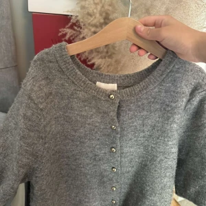 Grå stickad kofta från H&M - Mysig grå stickad kofta från H&M med korta ärmar och guldknappar knappar framtill. Perfekt att slänga på sig över en topp eller klänning. Enkel och stilren design som passar till mycket.