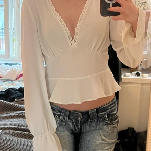 Vit blus med spetsdetaljer och volang - Säljer en söt vit blus med v-ringning och spetskant. Blusen har långa puffärmar med resår vid ärmslut och en volang nertill som ger en feminin touch. Perfekt att styla med jeans eller kjol.