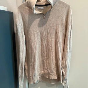 Beige stickad tröja med dragkedja från Jack & Jones - Säljer en beige stickad tröja från Jack & Jones med halv dragkedja vid halsen. Tröjan har lång ärm, ribbade muddar och en klassisk passform. Perfekt för lager-på-lager och enkel att matcha med olika outfits.