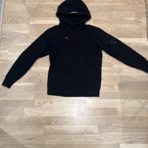 Svart hoodie från C.P. Company - Snygg svart hoodie från C.P. Company med en cool detalj på ärmen. Perfekt för en stilren look. Den har en klassisk huva med dragsko och en praktisk ficka framtill. Säljer den för den är lite förliten för mig. Frågor eller funderingar är det bara att slå en pling.
