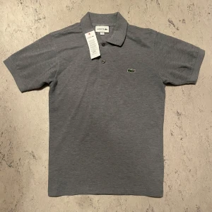 Lacoste Piké - Grå Lacoste piké i Xs, passar någon som är runt 170 beroende på vikt. Helt oanvänd utan några defekter.