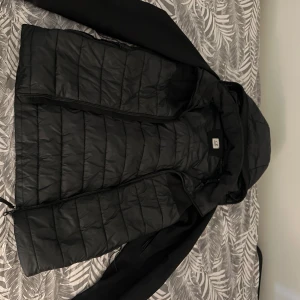 Svart pufferjacka från C.P. Company - Säljer en svart pufferjacka från C.P. Company med huva och dragkedja. Jackan har quiltad framsida och är tillverkad i ett mjukt, stretchigt syntetmaterial. Perfekt för kyligare dagar.