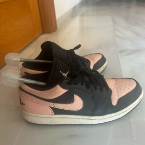 Nike Air Jordan 1 Low rosa och svarta sneakers - Säljer ett par Nike Air Jordan 1 Low sneakers i rosa och svart. Skorna har klassisk låg siluett, svart snörning och vit sula. Ikonisk Air Jordan-logga på hälen och swoosh i rosa på sidan. Perfekta för dig som gillar streetwear och snygga färgkombos.