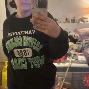 Svart sweatshirt med tryck från SHEIN - Svart sweatshirt från SHEIN med stort grönt och vitt tryck på bröstet: 'Vancouver British Columbia 1886 West Coast'. Tröjan har rund hals och långa ärmar. Perfekt för en avslappnad stil.
