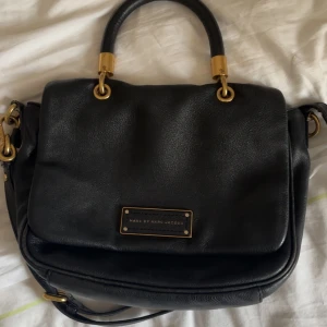 Svart axelväska från Marc by Marc Jacobs - Säljer en klassisk svart axelväska från Marc by Marc Jacobs med guldfärgade metalldetaljer och logoplatta framtill. Väskan har både handtag och justerbar axelrem, perfekt för att bära på olika sätt. Tillverkad i skinn med stilren design. Vintage, hjort ett extra hål i axelremmen så man kan ha den som kort Schulder bag 