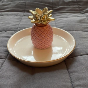 Smyckesfat med ananas i keramik - Superfint smyckesfat i form av en ananas. Fatet är vitt med en rosa ananas i mitten och guldfärgad topp. Perfekt för att förvara ringar, armband eller andra småsaker. Snygg detalj till rummet!
