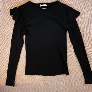 Svart långärmad topp med volang från Reserved - Säljer en svart långärmad topp från Reserved. Toppen har volangdetaljer på axlarna som ger en snygg touch. Mjuk och stretchig i materialet, perfekt till många olika outfits.