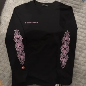 Svart långärmad tröja från Chrome Hearts - Svart långärmad tröja från Chrome Hearts med rosa tryck på bröstet, ryggen och längs båda ärmarna. Tröjan har unika grafiska detaljer och logga både fram och bak. Perfekt för dig som gillar streetwear och vill sticka ut.