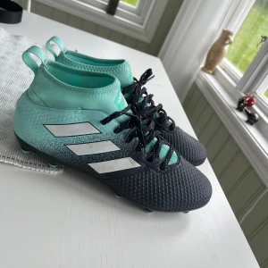 Adidas fotbollsskor i turkos och svart - Säljer ett par Adidas fotbollsskor i turkos och svart med vita ränder på sidan. Skorna har snörning och en högre strumpliknande krage för extra stöd. Perfekta för dig som vill sticka ut på planen!