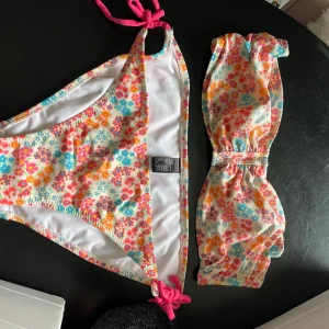 Blommig bikini med knytband - Söt bikini med blommigt mönster i rosa, orange, blått och gult. Överdelen är bandeau-modell med knyt i ryggen och underdelen har knyt i sidorna. Perfekt för sommardagar på stranden!